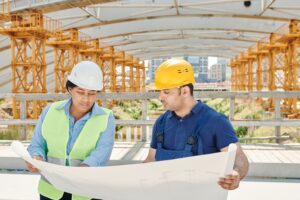 Two construction workers in hard hats review blueprints on a jobsite covered by performance bonds for government contracts.
